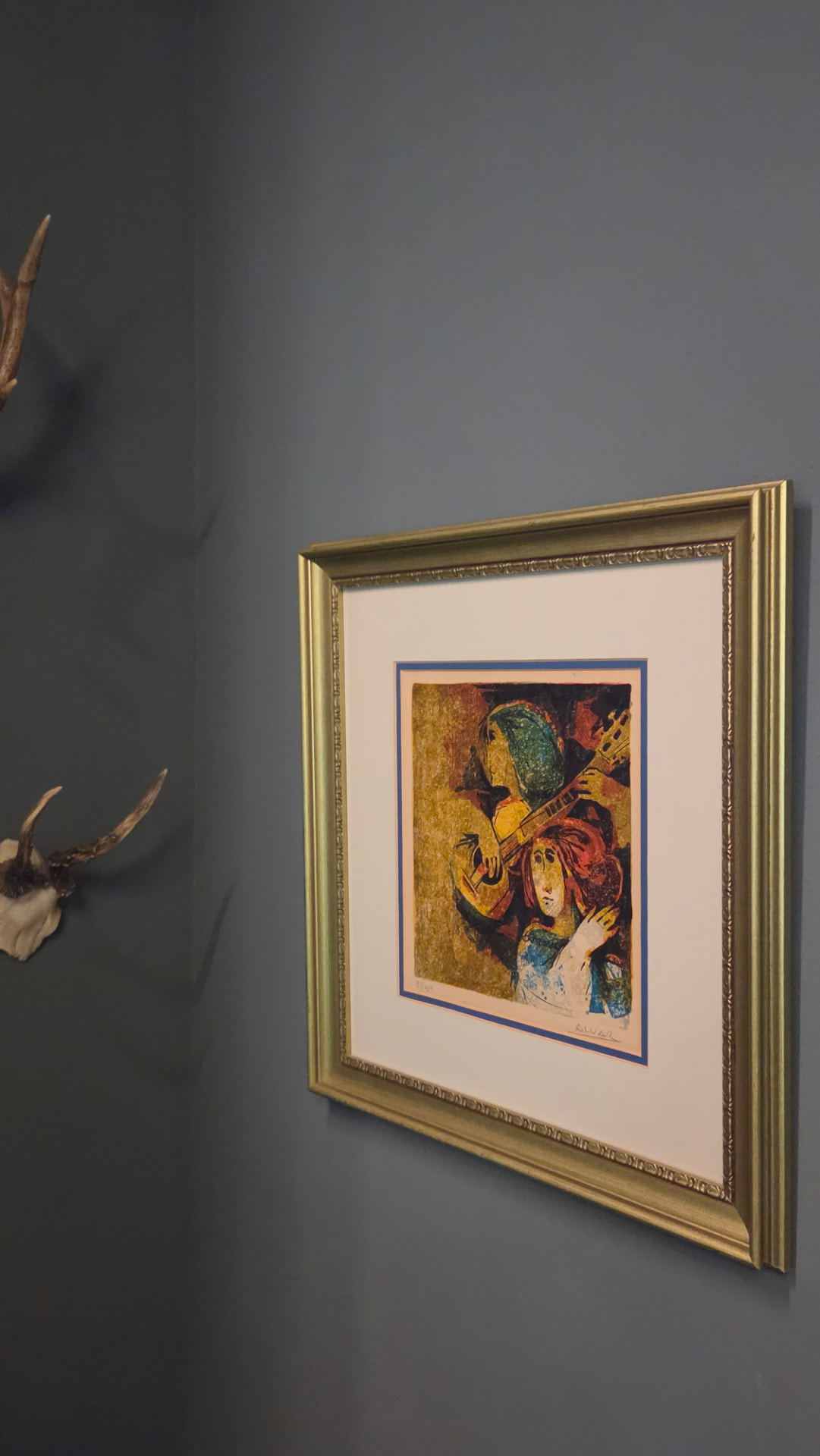 A modernist print of a guitar player in a gilt frame beside an antler mount on a slate green wall