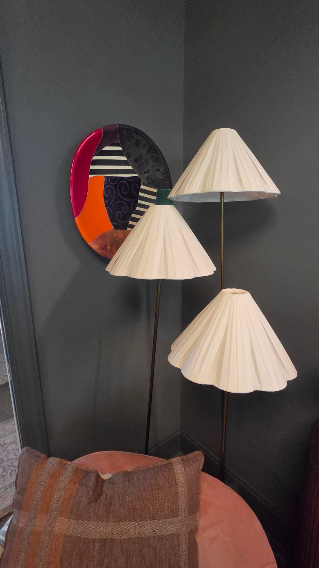 An oval hand-painted ceramic art plate hung between two pleated floor lampshades above a pink velvet chair