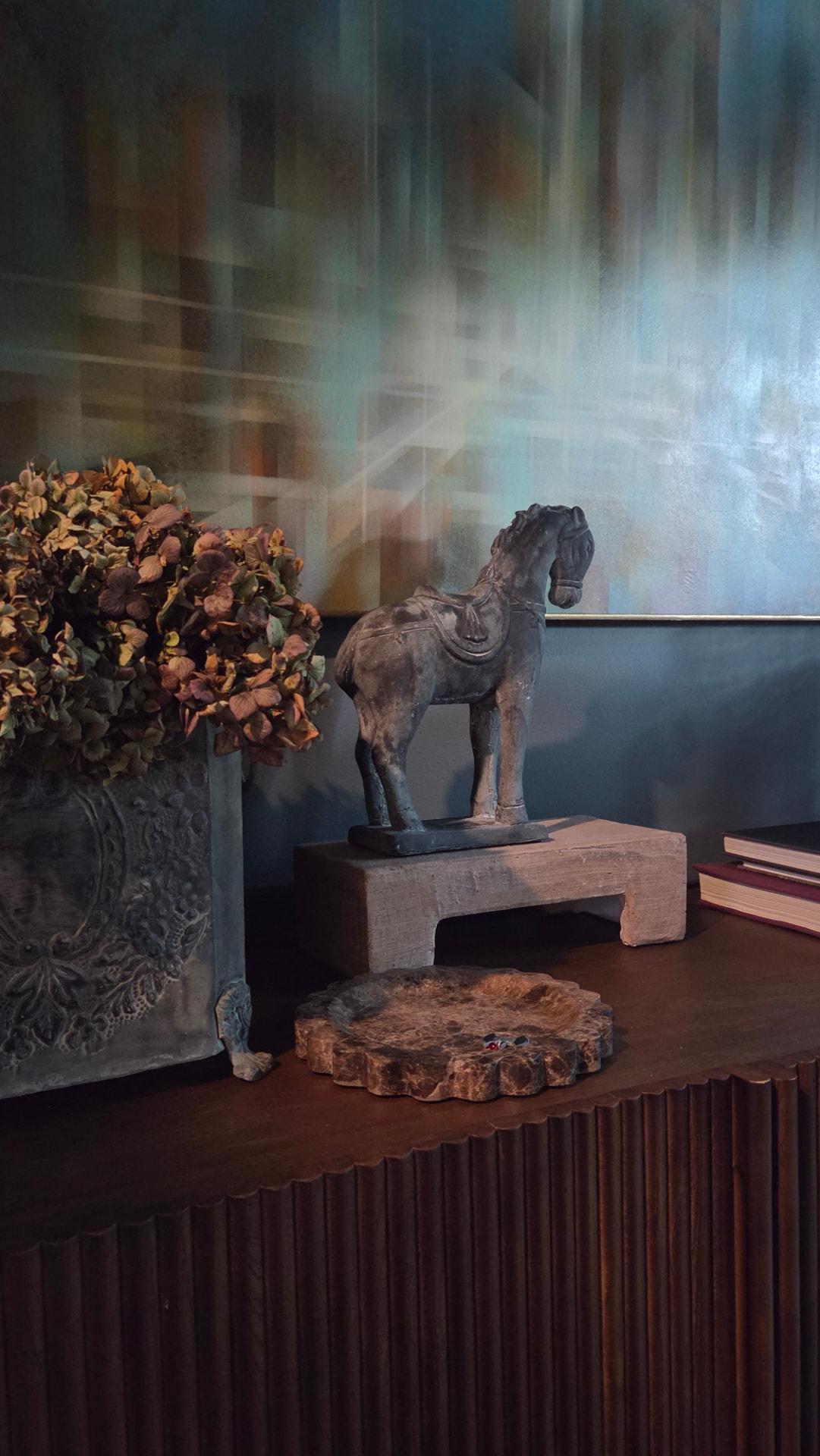 A carved Tang-style horse sculpture on a stone plinth beside dried hydrangeas, beneath a large abstract oil painting